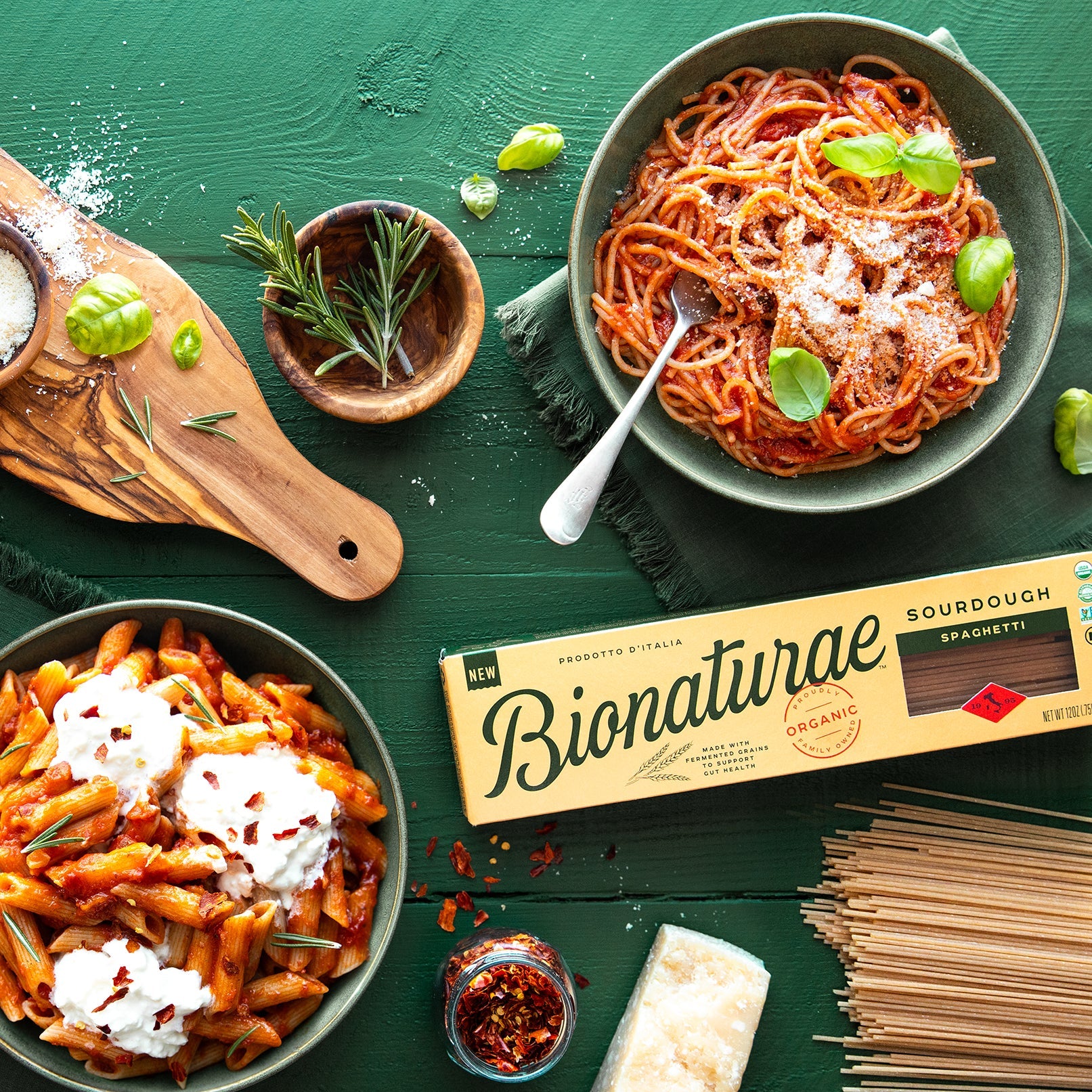 Green bowls with pasta and tomato sauce on a green background next to a box of Bionaturae pasta, fresh herbs and cheese