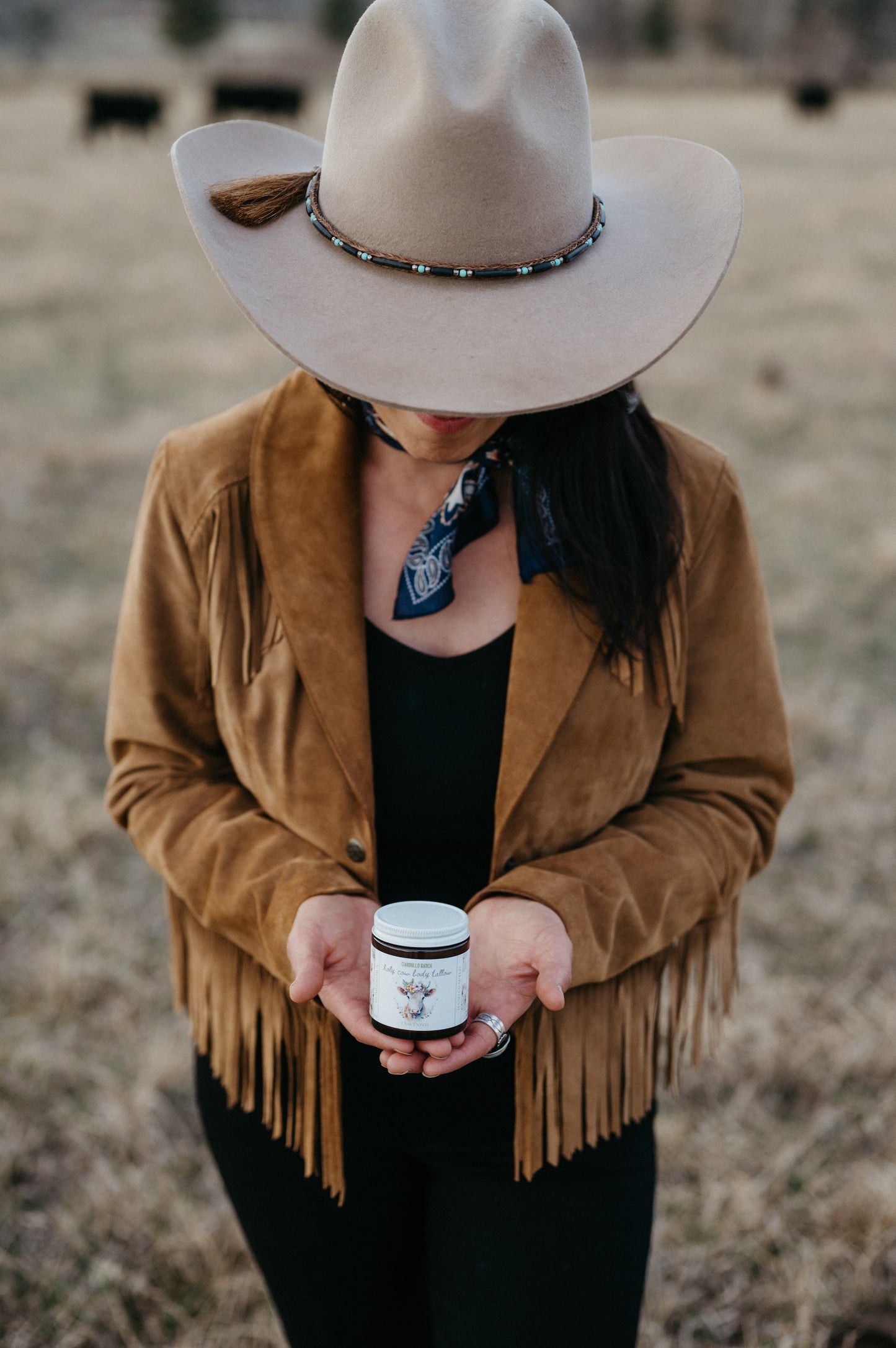 Woman wearing a cowboy hat and brown jacket holding a jar of tallow standing on a regenerative farm