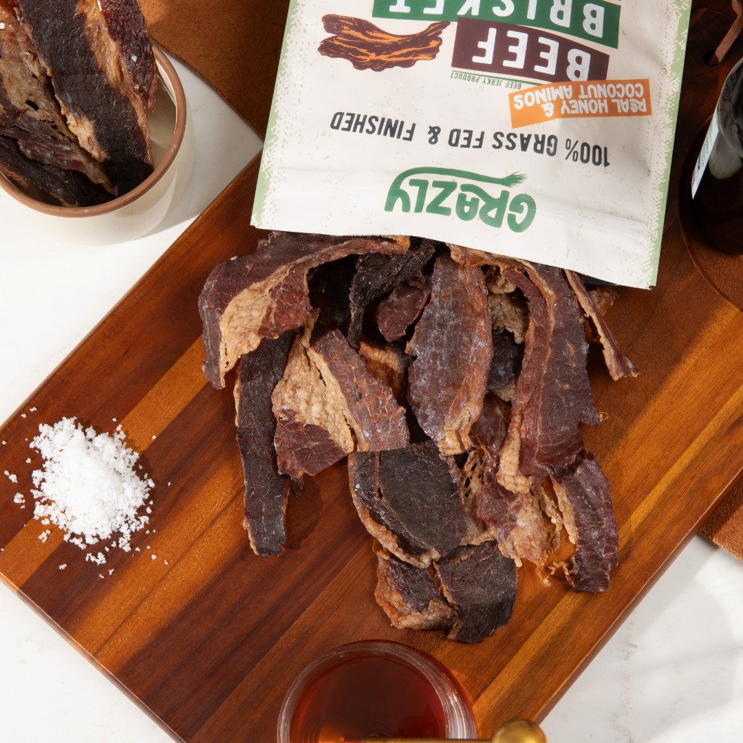 Dried beef jerky on a wooden board with a package labeled 'Grazly' in the background.