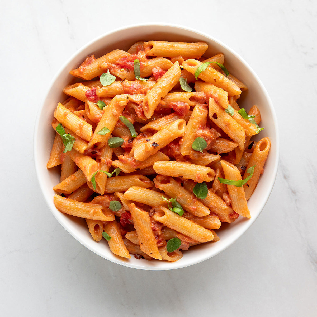 White bowl of penne tossed in tomato sauce, topped with micro herbs on a marble background